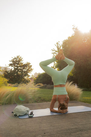Young woman in sportswear standing on her head on fitness mat at public park, doing yoga. Healthy...