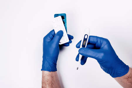 Man hands in blue disposal gloves hold thermometer and medicine pills on whiteの写真素材