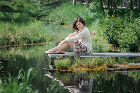 Portrait of a beautiful girl in Slavic clothes. Young barefoot woman is sitting by the pond. Summertimeの写真素材