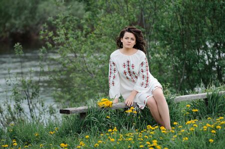 Portrait of a beautiful girl in Slavic clothes. Young woman with a bouquet of dandelions sitting on a wooden bench on the river bank. Summertimeの写真素材