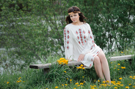 Portrait of a beautiful girl in Slavic clothes. Young woman with a bouquet of dandelions sitting on a wooden bench. Summertimeの写真素材