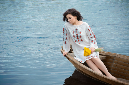 Portrait of a beautiful girl in Slavic clothes. Young woman with a bouquet of dandelions is sitting in a wooden boat. Summertimeの写真素材