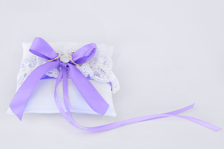 Two gold wedding rings on a white pillow with a purple bowの写真素材