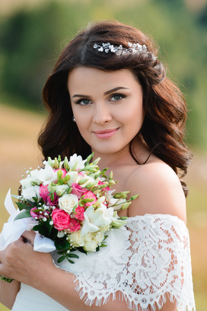 Beautiful brunette girl with a bouquet of flowers. Young woman with long hair in a white lace dress. Summer female portraitの写真素材