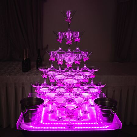 Pyramid of glasses for beverages, wine, champagne. Festive decoration for event, wedding, party. Wine glasses in neon lightの写真素材