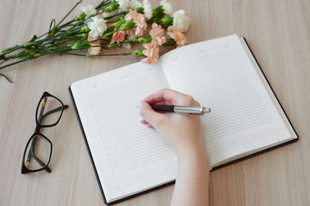 Female hand writing notes in daily planner. Open daily planner, bouquet of carnation flowers,...