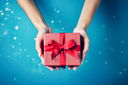 Female hands holding a gift box with a red bow on a blue background.の素材