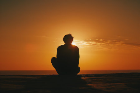 A man sits on the beach at sunset, looking out at the ocean. The sky is a beautiful mix of orange and pink hues, creating a serene and peaceful atmosphere. The man is lost in thoughtの素材