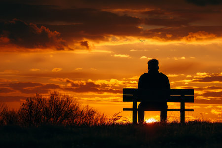 A man sits on a bench in the evening, watching the sun set. The sky is filled with clouds, creating a moody atmosphere. The man is lost in thoughtの素材