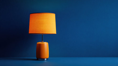 A bright orange lamp with a textured shade is positioned on a smooth surface, creating a striking contrast against a dark blue background, adding a pop of color to the room.の素材