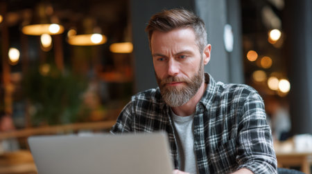 Man with a beard is intently typing on his laptop at a coffee shop surrounded by warm lights.の素材