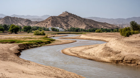 A river flows gently through a dry and sandy landscape, flanked by small green trees and hills. In the distance, a building sits atop a hill under clear skies.の素材