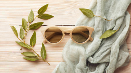 Sunglasses lie among soft fabric and fresh leaves on a serene wooden surface evoking relaxation.の素材