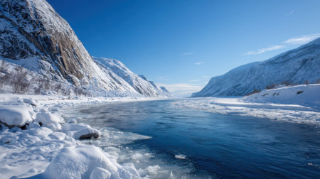 A tranquil winter scene showcases a river surrounded by tall mountains and covered in snow. The bright blue sky enhances the peaceful atmosphere of this beautiful outdoors.の素材