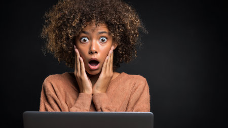 A young woman with curly hair reacts in shock while looking at her computer screen in a dark setting.の素材