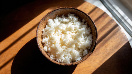 Fluffy white rice fills a rustic bowl illuminated by warm sunlight streaming through a window.の素材