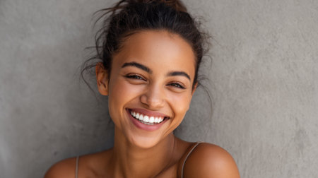 Joyful young woman shares a warm smile showcasing her natural beauty in a relaxed environment with a simple textured wall. The soft light enhances her cheerful expression.の素材