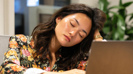 A woman with dark hair is resting her head on her hand at a desk. She appears tired, surrounded by a contemporary office backdrop and greenery. The warm lighting adds a calm atmosphere.の素材
