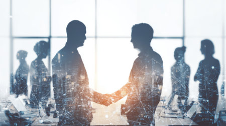 Two business professionals engage in a handshake during a meeting in a well-lit office. Silhouettes of other colleagues are present in the background, creating a collaborative atmosphere.の素材