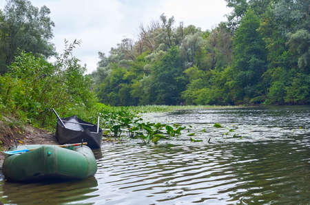 A small fishing inflatable rubber dinghy and metal boat with oars along river shore on summer rainny day. Summer landscape with river flow, boats, greenery and cloudy skyの写真素材