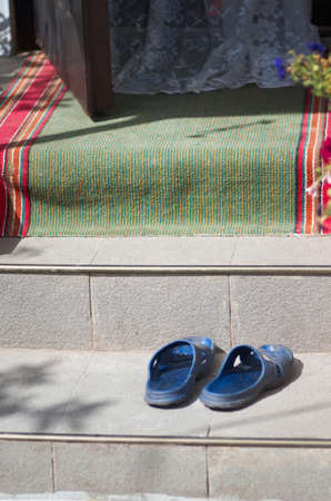 Blue sliders on steps of a house with open door, curtain lace and green-red footclothの写真素材