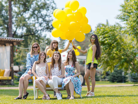 Group of beautiful young female friends posing outdoor with yellow balloons.Young women sitting on a bench and having funの写真素材