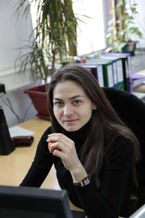 Portrait of a young smiling business woman in office.She holding an orange in the hand and working with the computer.Working place,office life, business conceptの写真素材