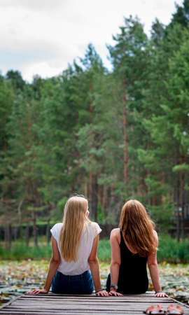 Two women are sitting together on pier enjoying the scenic view of a lake,back view. Female best friends enjoying a day.Beautiful romantic sunset imageの写真素材