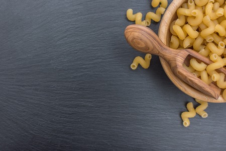 Pasta makaroni and olive wood scoop in a wooden bowl on black background of slate or stone Top view. Free space for your textの写真素材