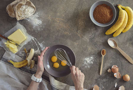 Banana bread baking concept, woman's hands are whisking eggs in a bowl, banana bread ingredients are lying on a kitchen table surface, view from above, space for a textの写真素材