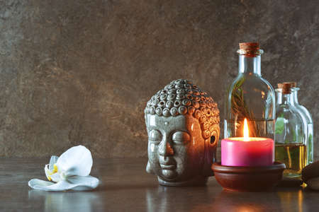 Natural spa cosmetic products such as massage infused oils arranged in a composition with birning candles and Buddha head statue, front view, space for a textの写真素材