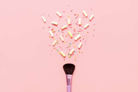 Make up concept background with a space for a text, cosmetic brush lying down on a pastel pink surface and marshmallows and confectionery candies are splashing out of the brushの写真素材