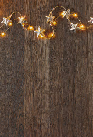 Christmas garland with golden stars lying down on wooden background lighten up by decorative lights, view from above, space for a greeting textの写真素材