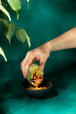 Vegan spring rolls concept, front view of a hand dipping a spring roll in a bowl with peanut sauce
