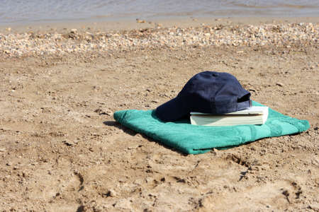blue baseball hat on green towel by the see and bookの写真素材