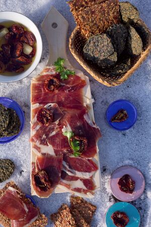 A cutting board  with  Traditional  tapas    with  spanish cured meat, bread chips with seed,  sun dried tomato with   garlic, parsley on background with snowの写真素材