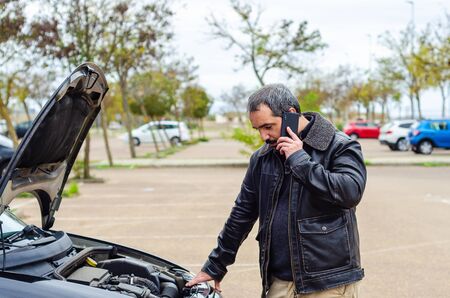 worried mature man looking under the hood of breakdown car and using smartphone