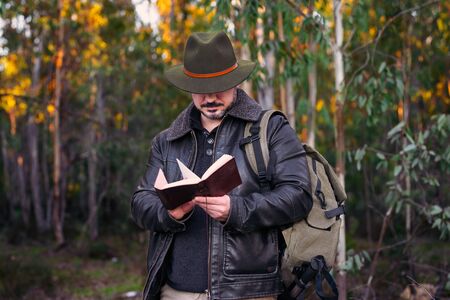 Mature man with hat and jacket looking old book on forest with copy space for text