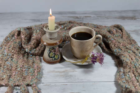 Composition in beige colors. Behind a cup of coffee is a warm knitted scarf. On the saucer are several flowers tied with a ribbon. Nearby is a candlestick with a burning candle.の写真素材