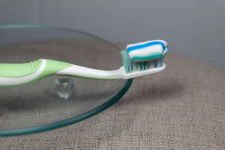 On a gray background on a transparent saucer lies a green toothbrush with toothpaste.