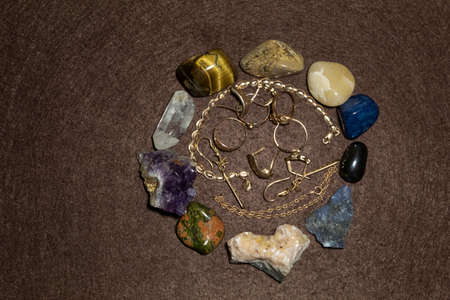View from above. On brown felt are gold womenâ€™s jewelry. Around are semiprecious stones.