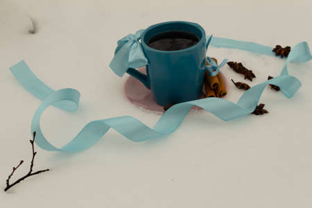 A blue cup with tea on a pink saucer stands on a white snow. Nearby are cinnamon sticks and star anise. Around lies a blue ribbon.の写真素材
