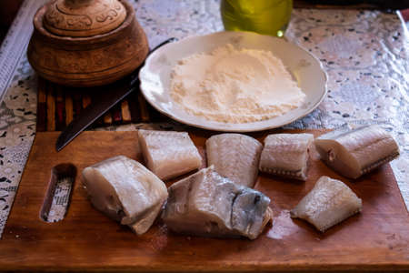 Pieces of fish lie on a cutting board on the table. In the background there is flour, oil and salt...