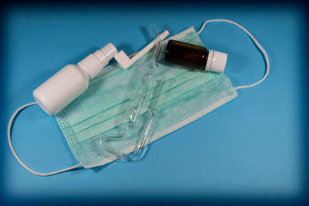 On a blue background lies a medical mask on which there is a spray for the throat, tea tree oil in a...