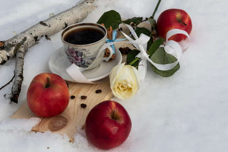 In the snow on a board is a cup of coffee. Nearby lies a white rose and three red apples.