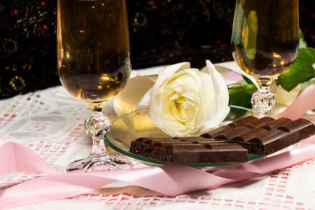 On the table there are two glasses of champagne lies a white rose and chocolate.の写真素材