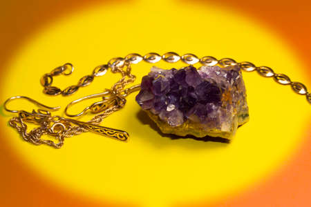 On a yellow background in an oval frame lies a semi-precious stone amethyst. Nearby are gold earrings and a bracelet.の写真素材