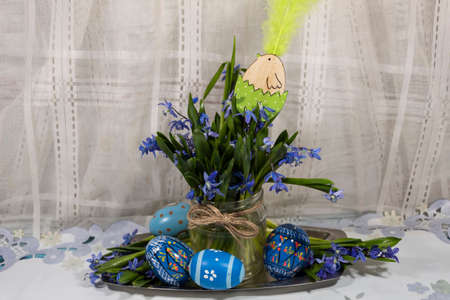 On a metal tray is a transparent jar with snowdrops and a decorative chicken. Nearby are easter...