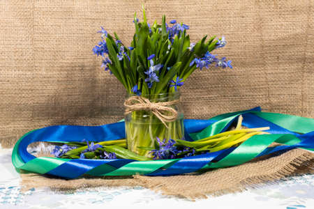 On a jute cloth on a metal tray is a jar with a bouquet of snowdrops. The composition is framed by a...