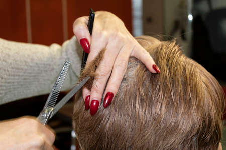 Hairdresser makes a short haircut to a woman. In her hands are special scissors and a comb.
